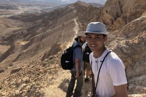 Two male Fellows stand on a trail in the desert, surrounded by mountains. One looks back at the camera and smiles.