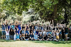 Group photo of over 40 Bronfman Fellows -- North American and Israeli -- who are sitting together on the grass under the trees, with their hands raised.