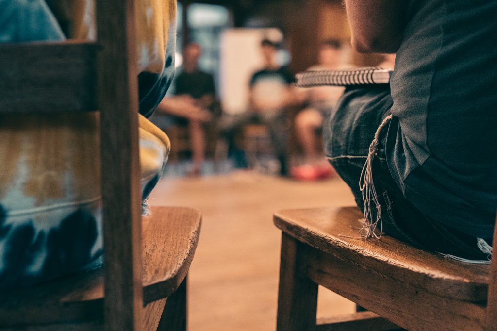 The backs of two Fellows sitting on wooden chairs. Tzitzit can be seen on one of them.